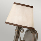 Introducing the Plymouth 1 Light Wall Light in bronze, a sophisticated, wall-mounted fixture with classic elegance. It features a beige fabric lampshade and a metal frame adorned with decorative scrollwork. The slightly angled shade beautifully complements the frame's muted brown finish, infusing any space with vintage charm.