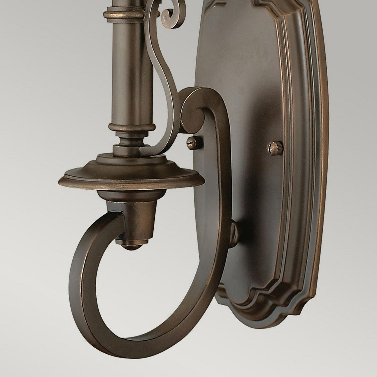 A close-up of the Plymouth 1 Light Wall Light - Bronze showcases classic elegance with its ornate, curved detailing and polished finish. The sconce includes a cylindrical base and a decorative backplate that add to its vintage charm. In the background, a plain light gray wall further enhances its timeless appeal.