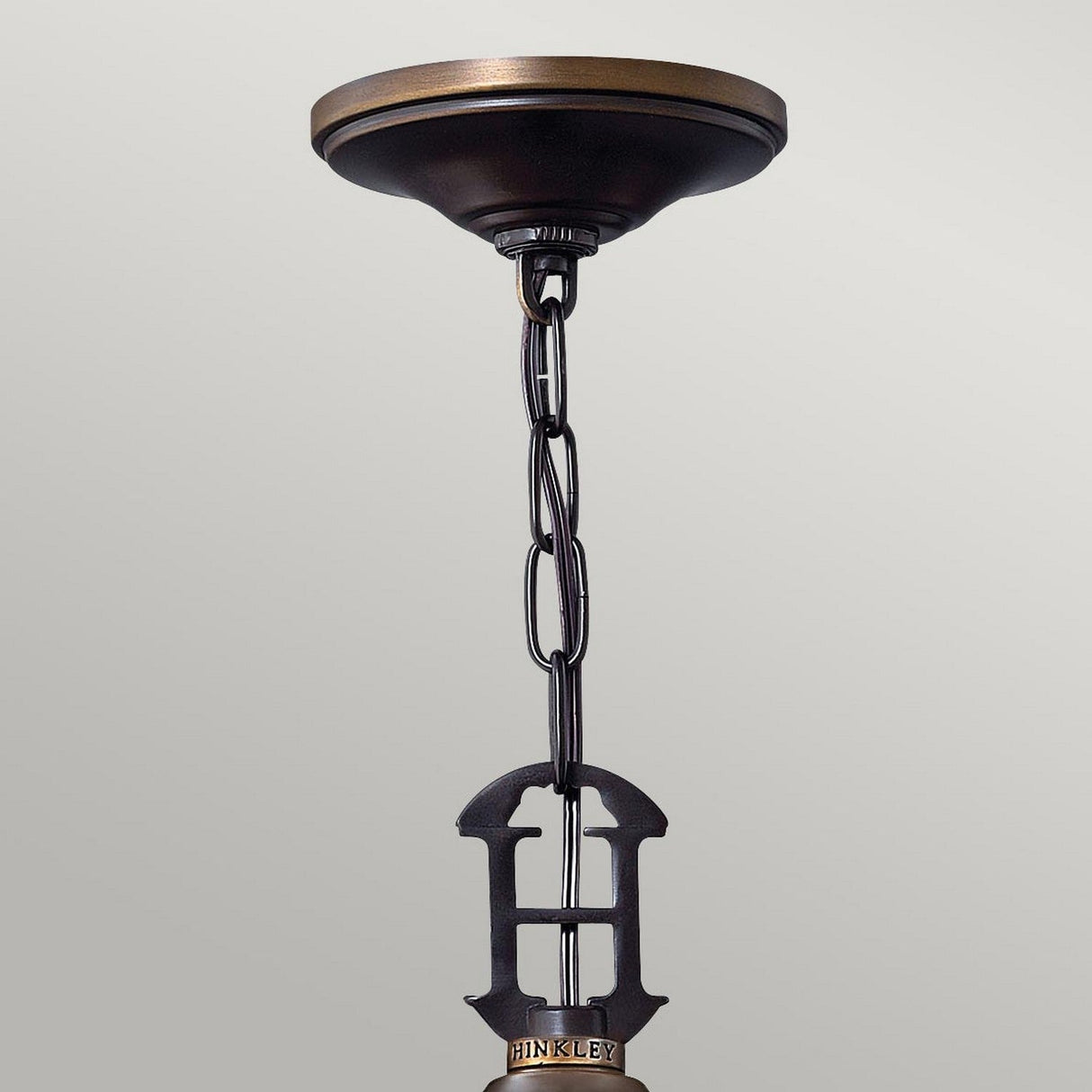 Close-up of a ceiling-mounted light fixture featuring a decorative chain and a metal component shaped like the letter H. The fixture showcases a vintage-inspired design, with the Plymouth 4 Light Chandelier in an Old Bronze finish mounted against a plain, light gray background.