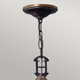 Close-up of a ceiling-mounted light fixture featuring a decorative chain and a metal component shaped like the letter H. The fixture showcases a vintage-inspired design, with the Plymouth 4 Light Chandelier in an Old Bronze finish mounted against a plain, light gray background.