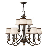 The Plymouth 9 Light Chandelier in Bronze is a large, elegant piece featuring an old bronze finish with nine cream-colored square lampshades arranged in two tiers. It boasts vintage-inspired candle sleeves, ornate curved arms, and decorative detailing, making it the perfect addition to classic or traditional interiors.