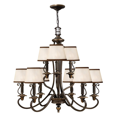 The Plymouth 9 Light Chandelier in Bronze is a large, elegant piece featuring an old bronze finish with nine cream-colored square lampshades arranged in two tiers. It boasts vintage-inspired candle sleeves, ornate curved arms, and decorative detailing, making it the perfect addition to classic or traditional interiors.