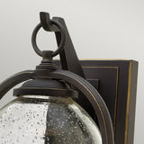 Close-up of the Quincy Outdoor 1 Light Medium Wall Lantern in Oil Rubbed Bronze, featuring a traditional design crafted from die-cast aluminum. It showcases a curved metal frame and a clear glass shade with a speckled texture, mounted on a rectangular base with decorative trim.