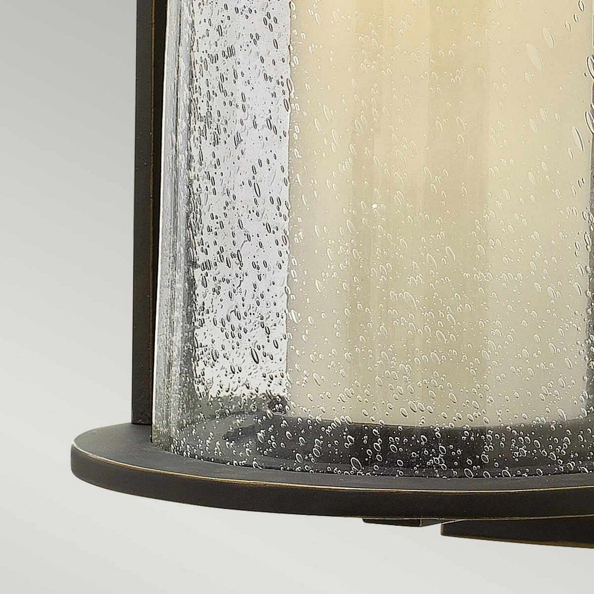 Close-up of the Quincy Outdoor 1 Light Medium Wall Lantern in Oil Rubbed Bronze, showcasing a lantern-style light fixture with a cylindrical frosted glass shade. The speckled glass beautifully complements the traditional design of the dark metal base, which may be crafted from die-cast aluminum, set against a light gradient background.