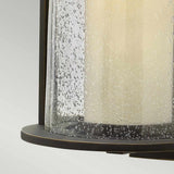 Close-up of the Quincy Outdoor 1 Light Medium Wall Lantern in Oil Rubbed Bronze, showcasing a lantern-style light fixture with a cylindrical frosted glass shade. The speckled glass beautifully complements the traditional design of the dark metal base, which may be crafted from die-cast aluminum, set against a light gradient background.