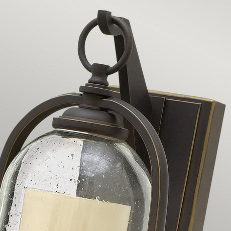 Close-up of the Quincy Outdoor 1 Light Small Wall Lantern in an oil-rubbed bronze finish. This lantern showcases a vintage aesthetic with a dark metal frame featuring an arched design and a clear glass shade speckled with small bubbles, all mounted on a rectangular backplate.