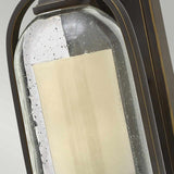 A close-up image showcases the Quincy Outdoor 1 Light Small Wall Lantern, which features a rectangular light encompassed by speckled glass adorned with water droplets. The lantern's metallic framework exhibits an elegant oil-rubbed bronze finish, and its intricate details are accentuated against a neutral background.