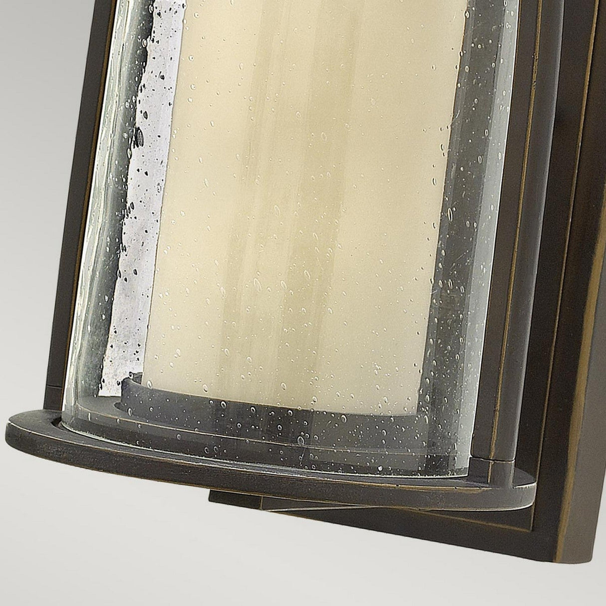 Close-up of the Quincy Outdoor 1 Light Small Wall Lantern, showcasing a cylindrical glass shade with tiny bubbles and an oil-rubbed bronze frame, set against a plain gray background.