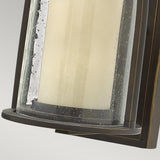 Close-up of the Quincy Outdoor 1 Light Small Wall Lantern, showcasing a cylindrical glass shade with tiny bubbles and an oil-rubbed bronze frame, set against a plain gray background.