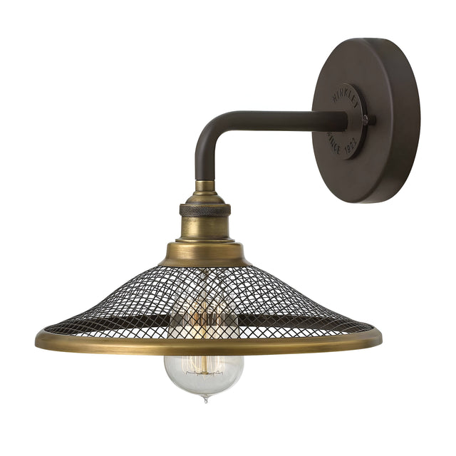 The Rigby 1 Light Wall Light in bronze and brass boasts a vintage design with a mesh shade and exposed bulb, mounted on a circular base. This industrial decor piece delivers an Americana aesthetic that channels the retro charm of barn lights.