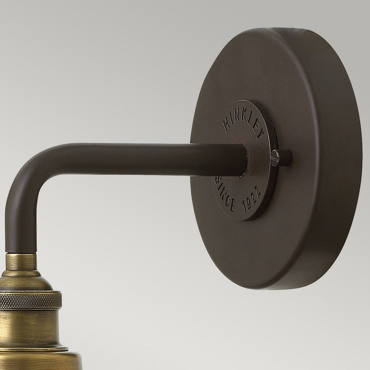 A close-up of the Rigby 1 Light Wall Light - Bronze & Brass shows a wall-mounted fixture with a round, dark metal base and a curved arm. The base is embossed with the text "Hinkley Since 1922." This barn light fixture exudes vintage Americana, combining a vintage brass finish against a light gray background.