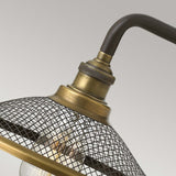 A close-up showcases the Rigby 1 Light Wall Light - Bronze & Brass, featuring an industrial-style lamp with a metal mesh shade and an exposed light bulb that embodies vintage Americana. Its brass-finished, angled arm evokes the charm of classic barn lights, all set against a plain light gray background.