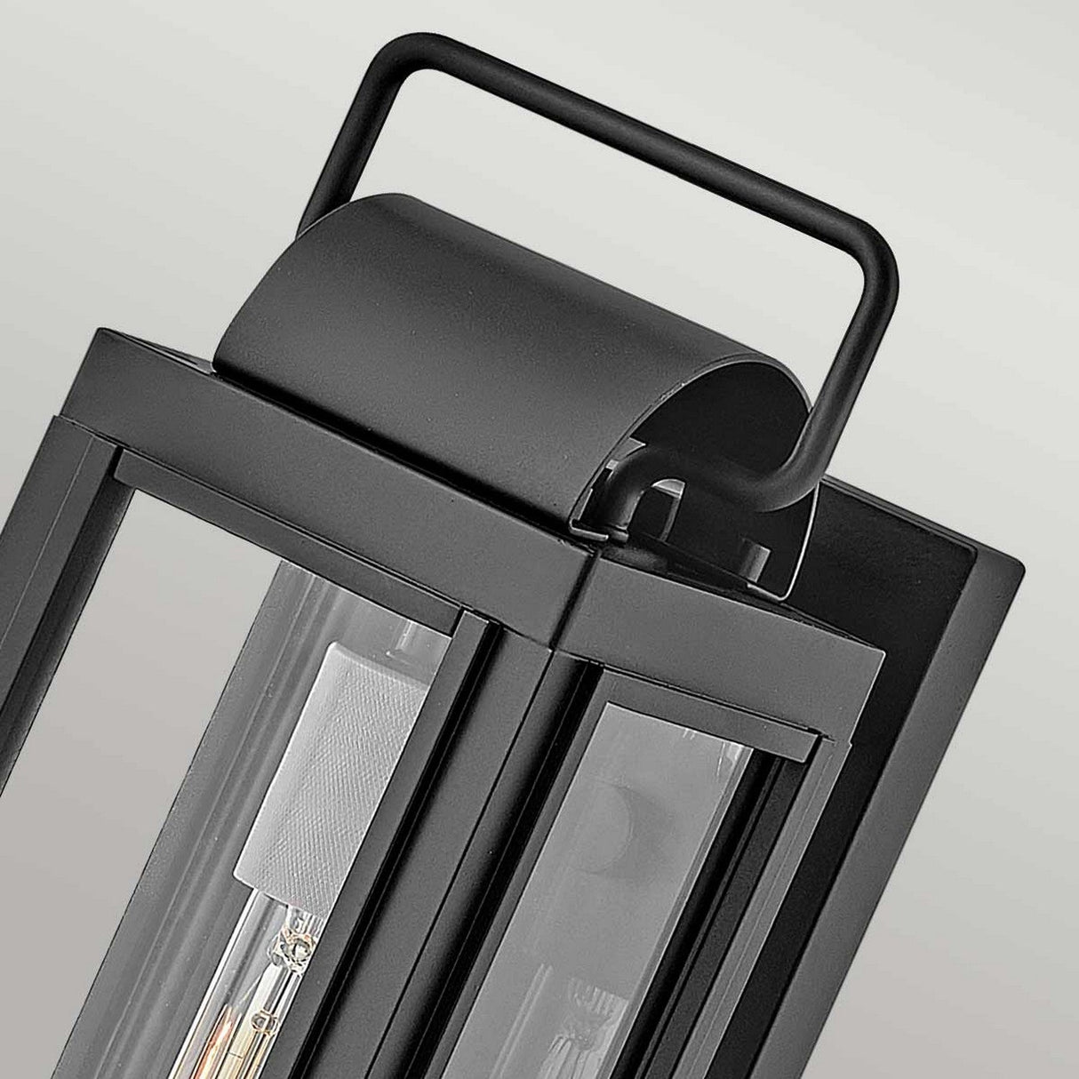 Close-up of the Sag Harbor Outdoor 1 Light Small Wall Lantern in Black, showcasing its contemporary design. Crafted from durable aluminum, it features a rectangular handle on top and glass panels that reveal the interior light bulb. The soft gray gradient background highlights its IP44-rated weather resistance.