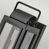 Close-up of the Sag Harbor Outdoor 1 Light Small Wall Lantern in Black, showcasing its contemporary design. Crafted from durable aluminum, it features a rectangular handle on top and glass panels that reveal the interior light bulb. The soft gray gradient background highlights its IP44-rated weather resistance.