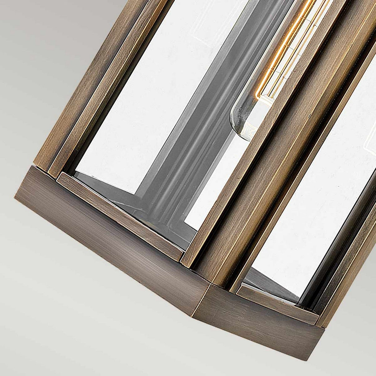 A close-up of the Sag Harbor Outdoor 1 Light Small Wall Lantern reveals its geometric pendant light fixture with a painted burnished bronze frame. It features clear glass panels that display an elongated filament bulb inside, embodying a modern and industrial design reminiscent of the product itself.