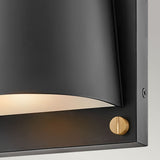 A close-up of the Scout Outdoor 1 Light Small Wall Light in a modern black finish showcases its curved, partially visible shade with a small brass accent screw at the bottom corner, mounted on a gray wall. The weather-resistant fixture emits a gentle illumination from within.