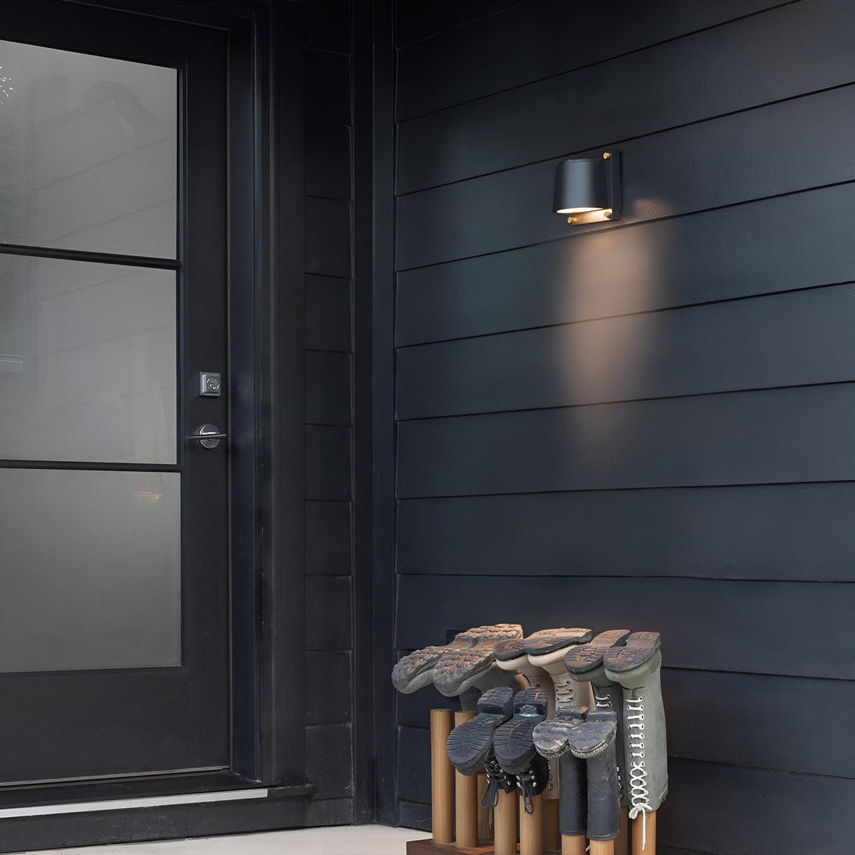 The sleek black exterior of the modern house features a closed door with a windowpane, illuminated by the Scout Outdoor 1 Light Small Wall Light in black. Below this weather-resistant fixture, a wooden shoe rack neatly holds nine pairs of boots in different styles and colors.