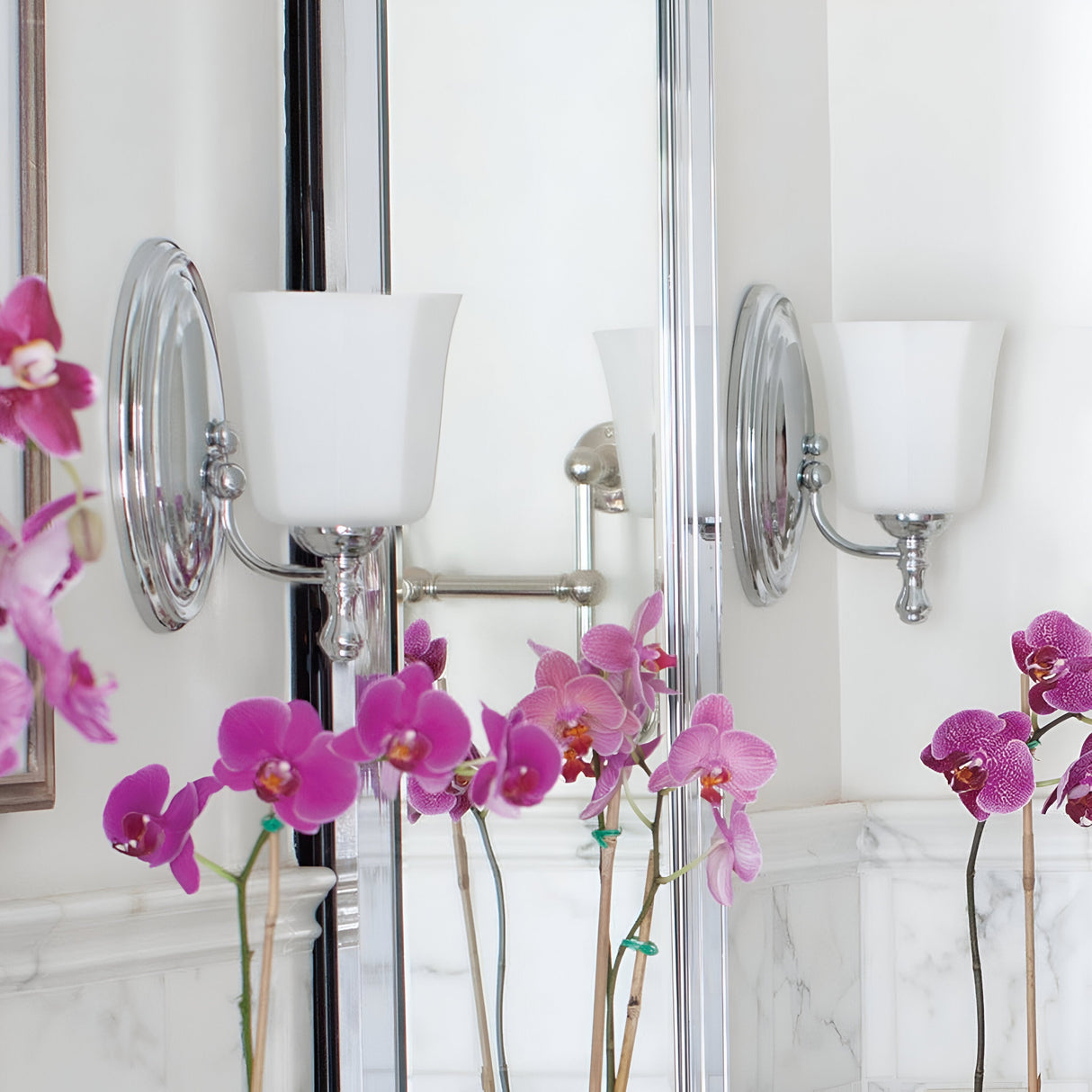 In the bathroom, the Shelly 1 Light Wall Light in polished chrome stands beside a mirror, adorned with a frosted glass sconce. Below, vibrant pink orchids beautifully contrast against a marble wall. The mirror captures the soft lighting and doubles the floral display, creating an elegant ambiance.