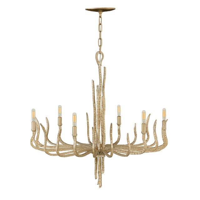 A Spyre 6 Light Chandelier in the Champagne Gold finish hangs from the ceiling with a rustic antler design. It features six light bulbs arranged in a circular pattern, emitting a warm glow. The chandelier has a natural, wood-like appearance accented with elegant Champagne Gold touches.