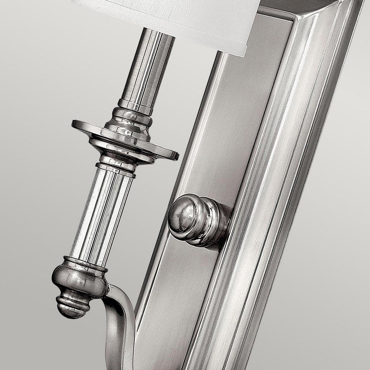 Close-up of the Sussex 1 Light Wall Light in brushed nickel, highlighting its intricate detailing and timeless elegance. The design includes a long vertical backplate and a cylindrical candle holder, partially visible against a light gray background, accentuating its sleek, silver finish.