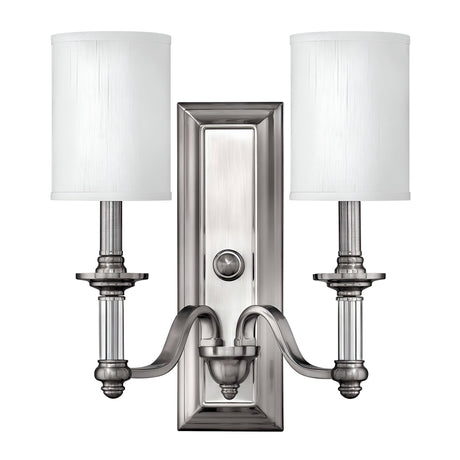 The Sussex 2 Light Wall Light - Brushed Nickel exudes contemporary elegance with its polished silver finish and white cylindrical lampshades. It is mounted on an ornate rectangular backplate, featuring curved arms and a central decorative knob in sleek Brushed Nickel, making it a perfect addition to any modern space.