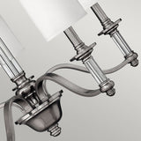 Close-up of the Sussex 5 Light Chandelier - Brushed Nickel, featuring metallic arms and cylindrical lampshades. Its brushed nickel finish exudes traditional charm with sleek curves and intricate detailing, offering a contemporary look against a neutral background.