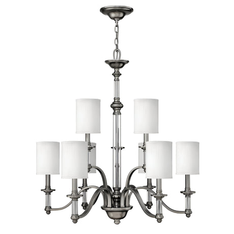 The Sussex 9 Light Chandelier in brushed nickel includes six cylindrical white fabric shades. It features a central stem and six elegantly curved arms extending outward, providing a symmetrical and stylish design ideal for any contemporary setting.