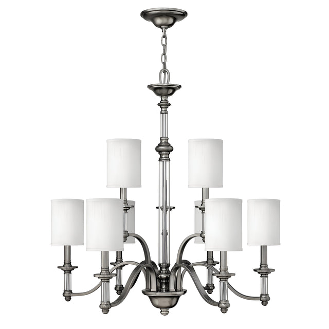The Sussex 9 Light Chandelier in brushed nickel includes six cylindrical white fabric shades. It features a central stem and six elegantly curved arms extending outward, providing a symmetrical and stylish design ideal for any contemporary setting.
