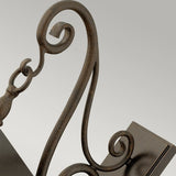 A close-up of the Trellis Outdoor 4 Light Large Wall Lantern in bronze reveals its ornate, dark metal scroll design with curved and intricate detailing set against a light gray background. This elegant wrought iron structure is reminiscent of an outdoor decorative fixture or piece of furniture.