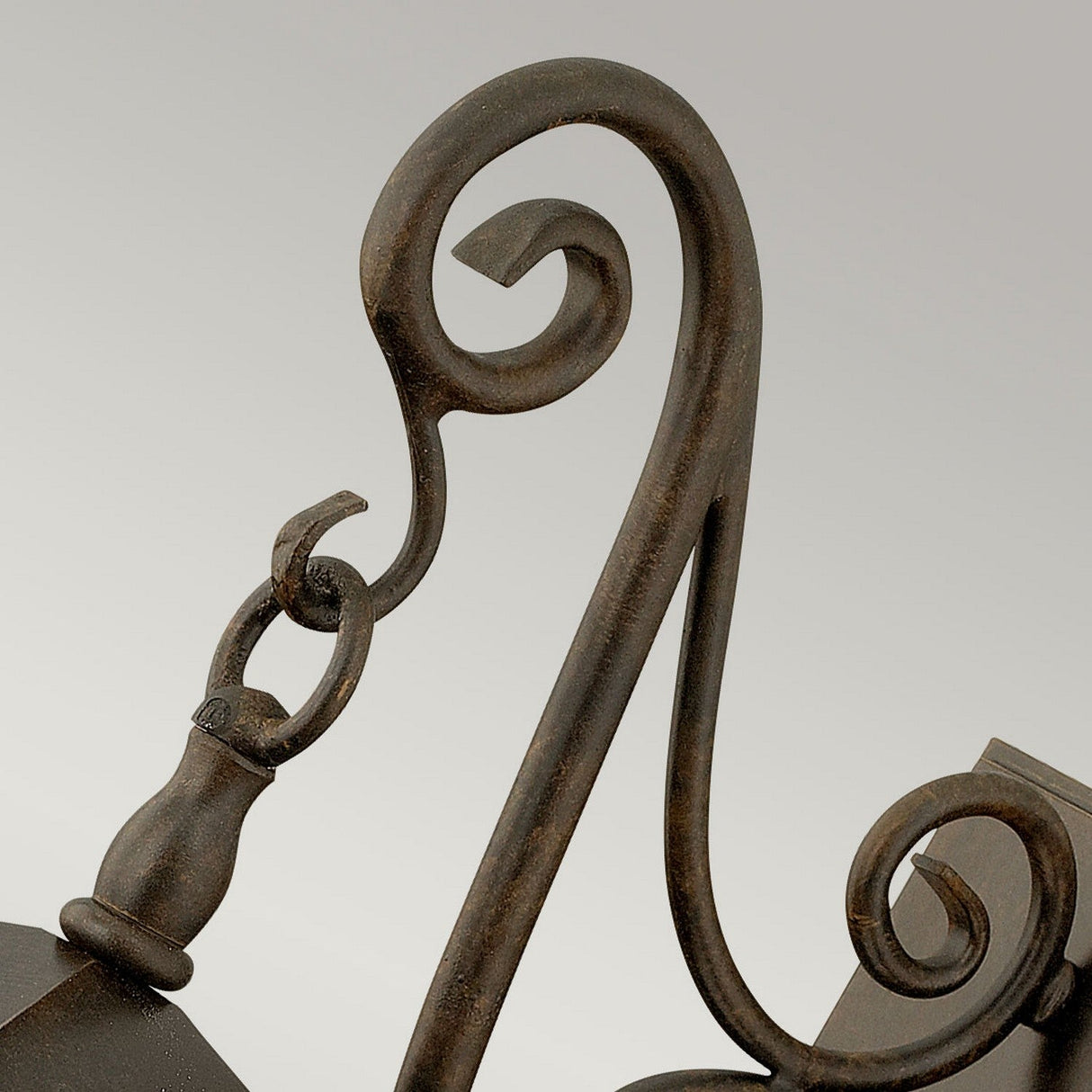 A close-up of the Trellis Outdoor 1 Light Small Wall Lantern in bronze showcases its ornate metal hook with a decorative scroll design, affixed to a rectangular bracket. The rustic, dark brown finish evokes the charm of vintage outdoor lighting, perfectly complemented by seedy glass panels against a plain, light gray background.