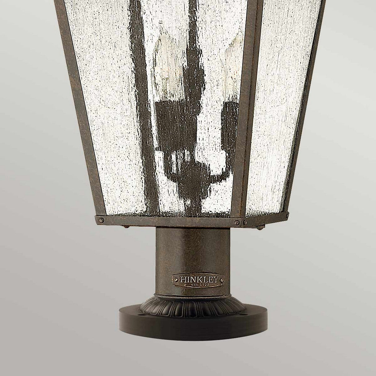 A close-up of the Trellis Outdoor 3 Light Large Pedestal in bronze showcases a textured glass design. This garden path illumination fixture is octagonal in shape and prominently displays the Hinkley brand on its base.