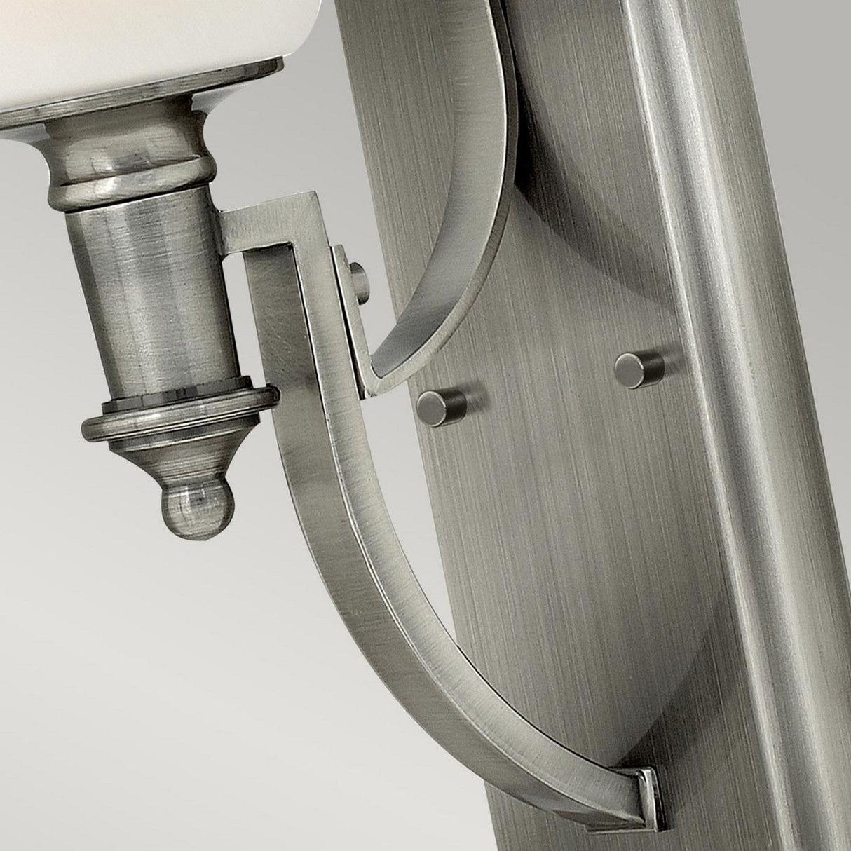 The Truman 1 Light Wall Light in antique nickel showcases a close-up of its brushed metal fixture, featuring a curved design with rivets. This sleek piece boasts an opal glass shade, offering a modern appearance with smooth finish and detailed craftsmanship.