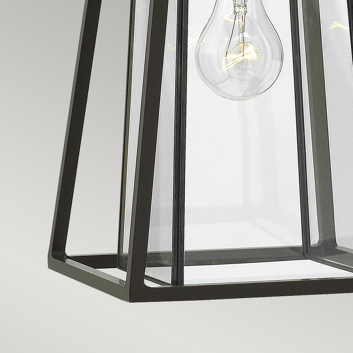 A close-up of the Walker Outdoor 1 Light Medium Wall Lantern, featuring a geometric metal frame in bronze with heritage brass, evokes the charm of rustic decor. Inside, a clear Edison bulb is visible, highlighting the lantern's contemporary design against a plain background.