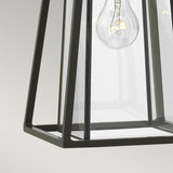 A close-up of the Walker Outdoor 1 Light Medium Wall Lantern, featuring a geometric metal frame in bronze with heritage brass, evokes the charm of rustic decor. Inside, a clear Edison bulb is visible, highlighting the lantern's contemporary design against a plain background.