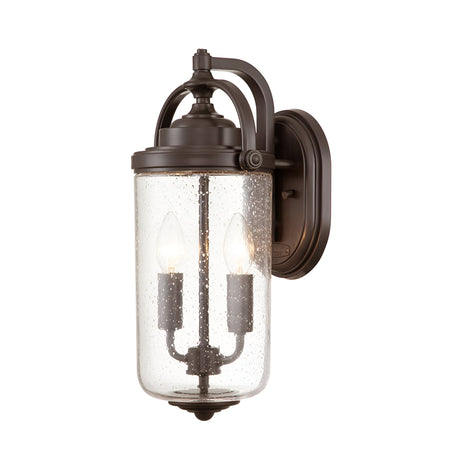 The Willoughby 2 Light Wall Lantern in Oil Rubbed Bronze features a cylindrical glass casing with a textured, speckled finish. Inside, two candle-shaped light bulbs shine through the clear seeded glass. This wall lantern is elegantly mounted on a circular plate.