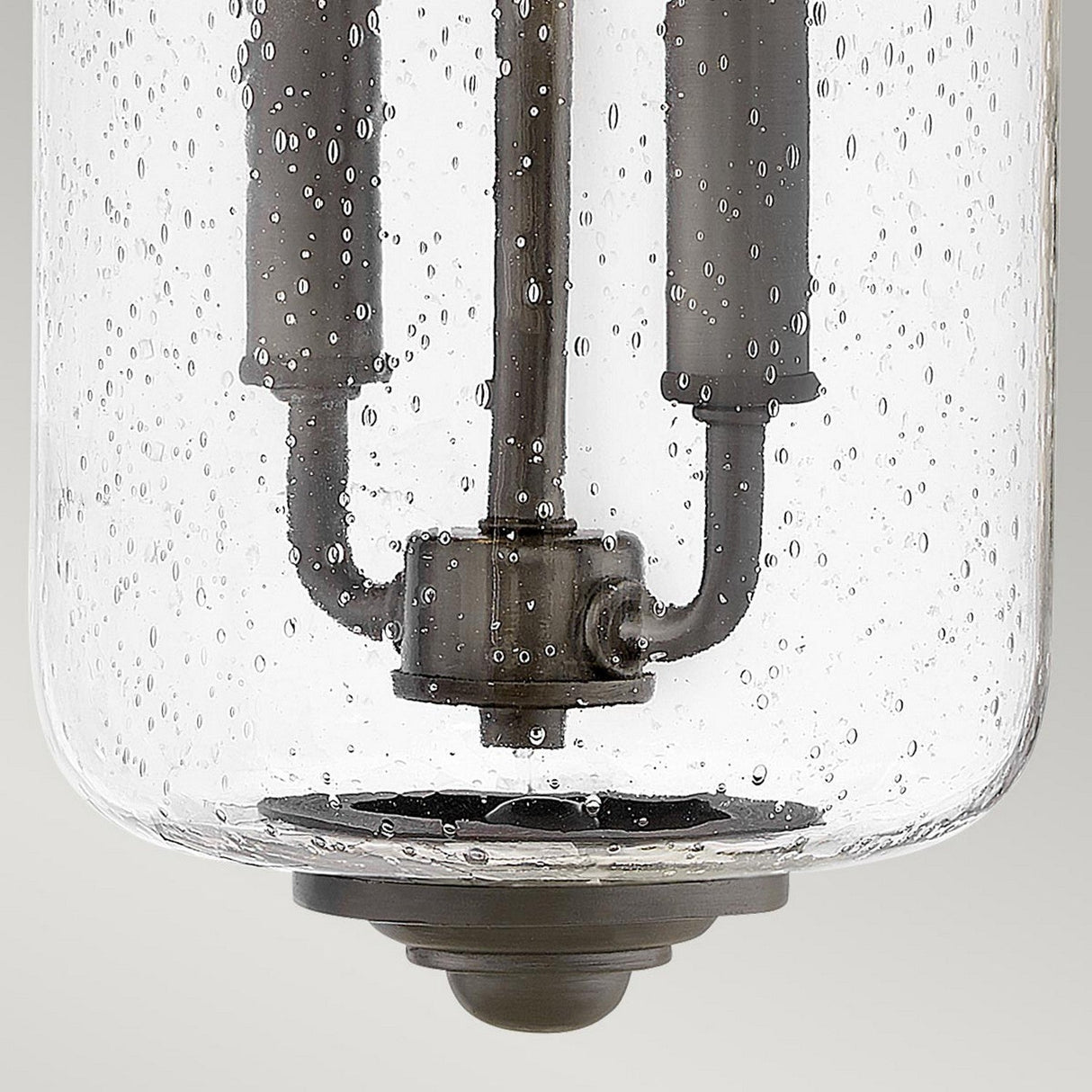 A close-up of the Willoughby 2 Light Wall Lantern - Oil Rubbed Bronze, showcasing its clear seeded glass cover adorned with air bubbles and revealing the metal components inside. The background remains plain and light-colored.