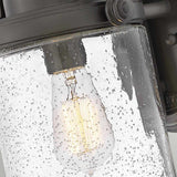 A close-up of the Willoughby 1 Light Wall Lantern reveals a vintage-style light bulb glowing inside a cylindrical glass fixture. The bubbled surface amplifies the warm, ambient glow. Constructed with oil rubbed bronze, this lantern infuses outdoor lighting with an industrial flair and dark metal charm.