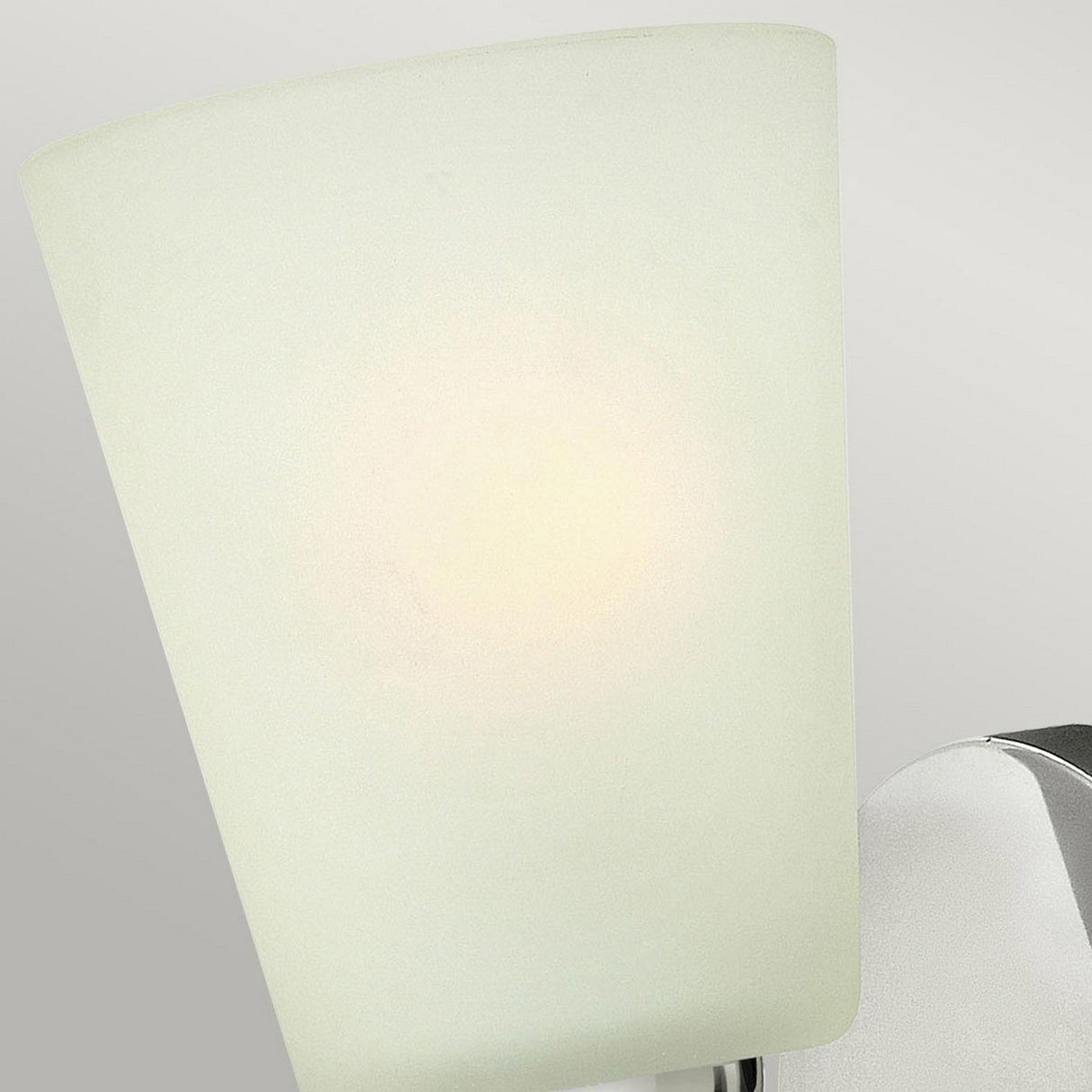 A close-up of the Zelda 1 Light Wall Light - Polished Nickel showcases a frosted glass lampshade affixed to its polished nickel fixture, exuding a mid-century vibe. Set against a light gray backdrop, it emits a soft, warm glow.