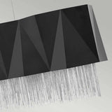 The Zuma 6 Light Linear Chandelier in Satin Black/Silver features a modern geometric design with a textured triangular pattern. It hangs elegantly against a light gray background, and its thin silver chains cascade down to create a fringe-like effect. This piece offers ample illumination and is ideal for enhancing contemporary interiors with its sleek style.