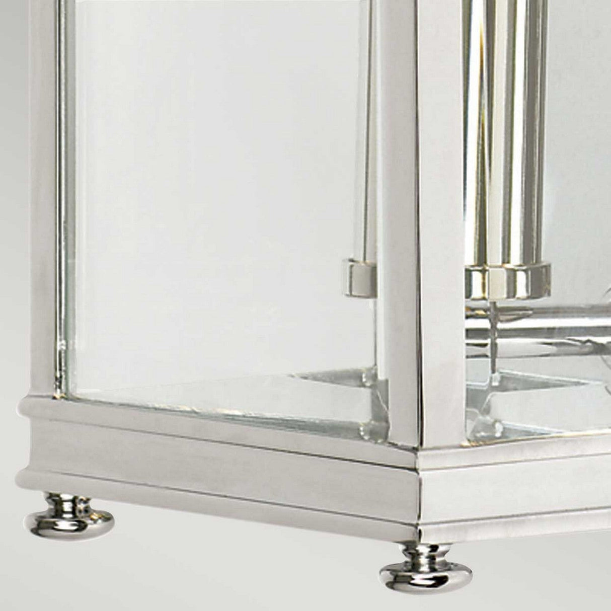 Close-up of the Holborn 1 Light Outdoor Small Wall Lantern in polished chrome, showcasing its modern rectangular glass and metal design inspired by Georgian lanterns. It stands on four small round feet, with a partially visible candle holder inside. The backdrop is an understated, neutral color.