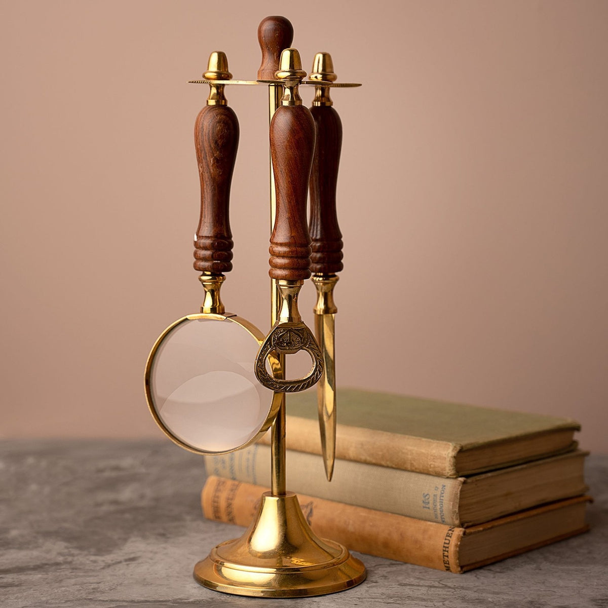 Desk tool stand in gold with brown turned handles holding a round magnifying glass, letter opener, and bottle opener, set beside stacked books on a grey surface.