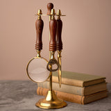 Desk tool stand in gold with brown turned handles holding a round magnifying glass, letter opener, and bottle opener, set beside stacked books on a grey surface.