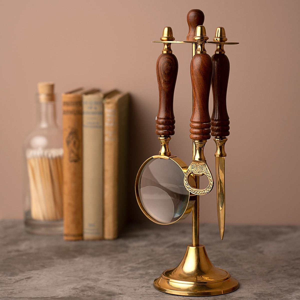 Desk stand holding a magnifying glass, letter opener, and bottle opener with brown turned wood handles and gold caps, set near books on a grey surface.