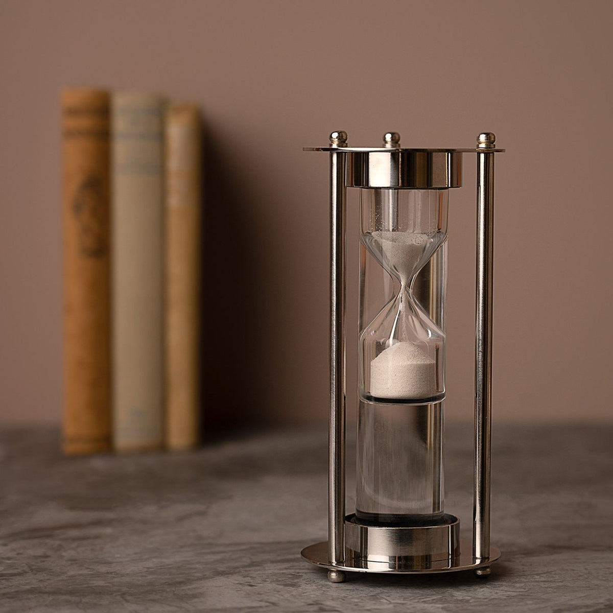 Water Timer Hourglass with Three Rods and Ball Feet in silver, white sand in a glass bulb framed by slim rods with ball finials, blurred books behind.