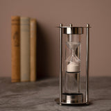Water Timer Hourglass with Three Rods and Ball Feet in silver, white sand in a glass bulb framed by slim rods with ball finials, blurred books behind.
