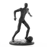 Black resin footballer figurine with textured surface, faceless head, and long limbs, mid-stride with one foot on a football, set on an oval base against a white background.