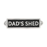 Rectangular metal wall plaque reading "DAD'S SHED" in raised silver capitals on a black panel with a silver border and two fixing holes, shown on a white background.