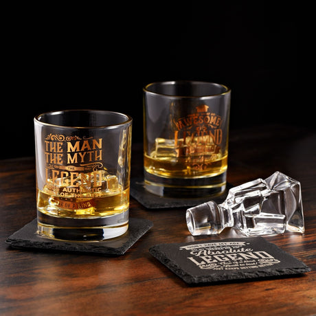 Clear glass tumbler with gold rim, reading "The Man The Myth The Legend" in gold lettering, filled with whisky and ice on a dark slate coaster.