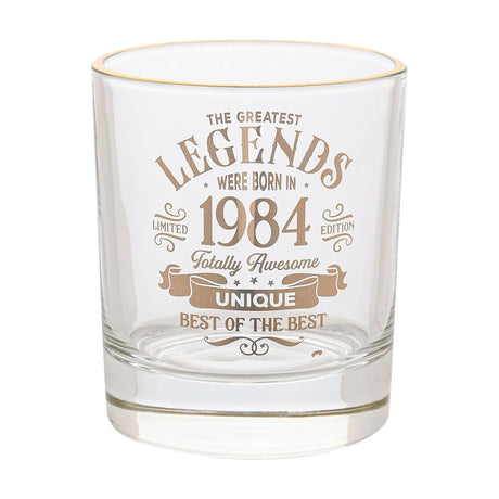 Clear glass whisky tumbler with a gold rim, printed in gold text "The Greatest Legends Were Born in 1984, Totally Awesome, Unique, Best of the Best," on a plain white background.