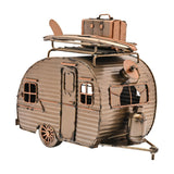 Gold metal teardrop caravan figurine with corrugated panels, door, framed windows, roof rack holding a surfboard and suitcase, and gas bottle, on a white background.